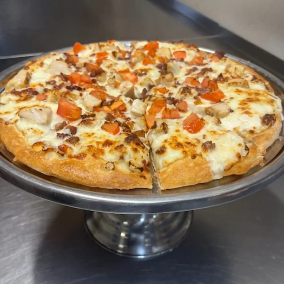 Cicero's Pizza with white sauce, chicken, bacon, and diced tomatoes on a metal stand.